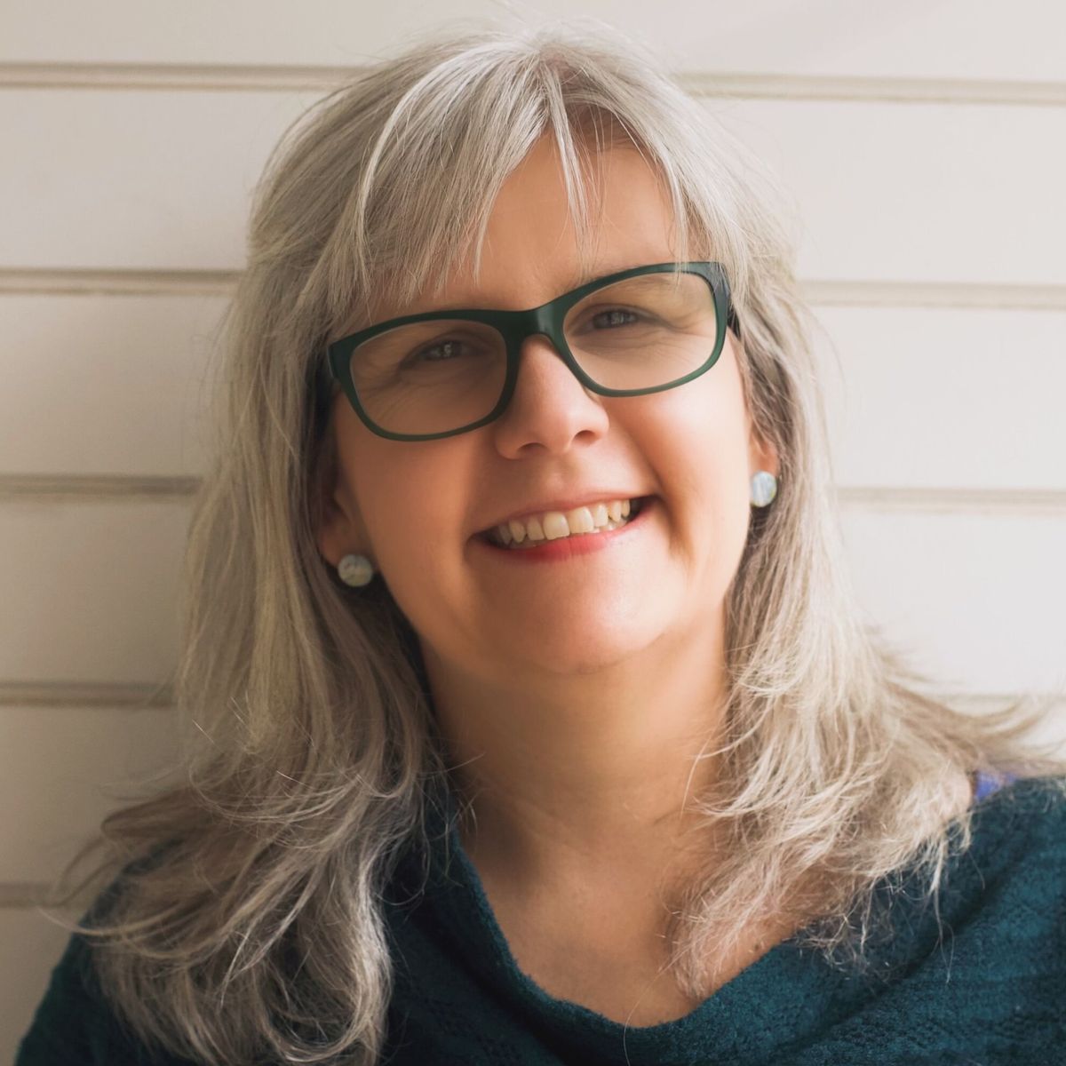 Headshot of Kate Robson, author and speaker. She is wearing glasses and a dark green knit sweater. She is standing in front of a white, panelled wall.